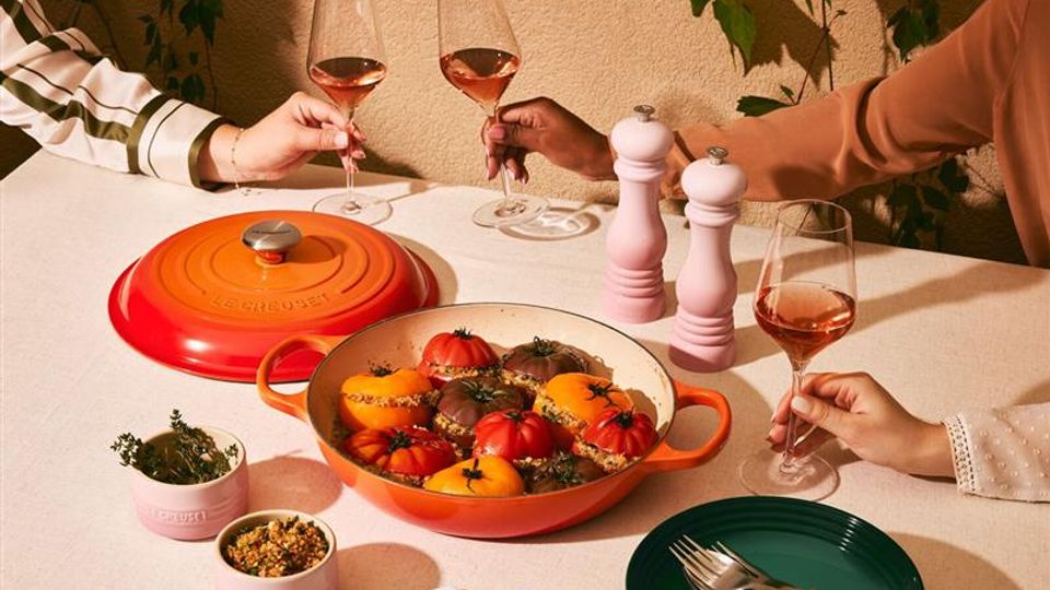Immagine di una pentola in ghisa Le Creuset di color arancione posta al centro di una tavola apparecchiata e contenente dei pomodori ripieni cucinati e serviti nella stessa